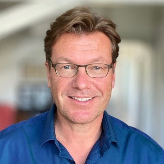 Porträt von Ben Buse , der in einem Innenraum steht und freundlich in die Kamera lächelt. Er trägt ein dunkelblaues Hemd.  