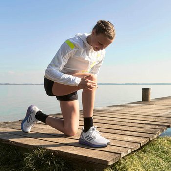 Das Bild zeigt Läuferin Anna Hahner, die am Ufer eines Sees auf einem Holzsteg ihr Knie untersucht. Erfahre, was du gegen Knieschmerzen beim Laufen unternehmen kannst.