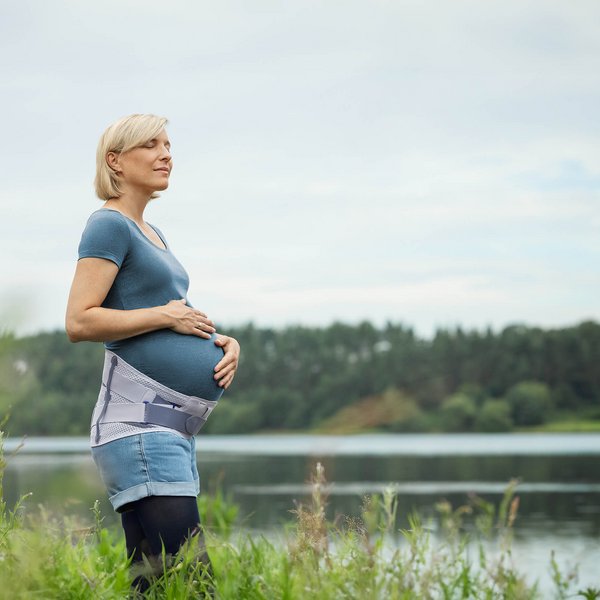 Schwangere Frau steht am Ufer eines Sees und hält ihren Babybauch, während sie in die Ferne blickt. Sie trägt die Spinova Mum Schwangerschaftsorthese und genießt die ruhige Naturkulisse.