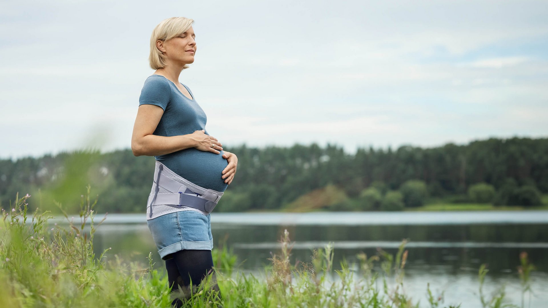 Schwangere Frau steht am Ufer eines Sees und hält ihren Babybauch, während sie in die Ferne blickt. Sie trägt die Spinova Mum Schwangerschaftsorthese und genießt die ruhige Naturkulisse.