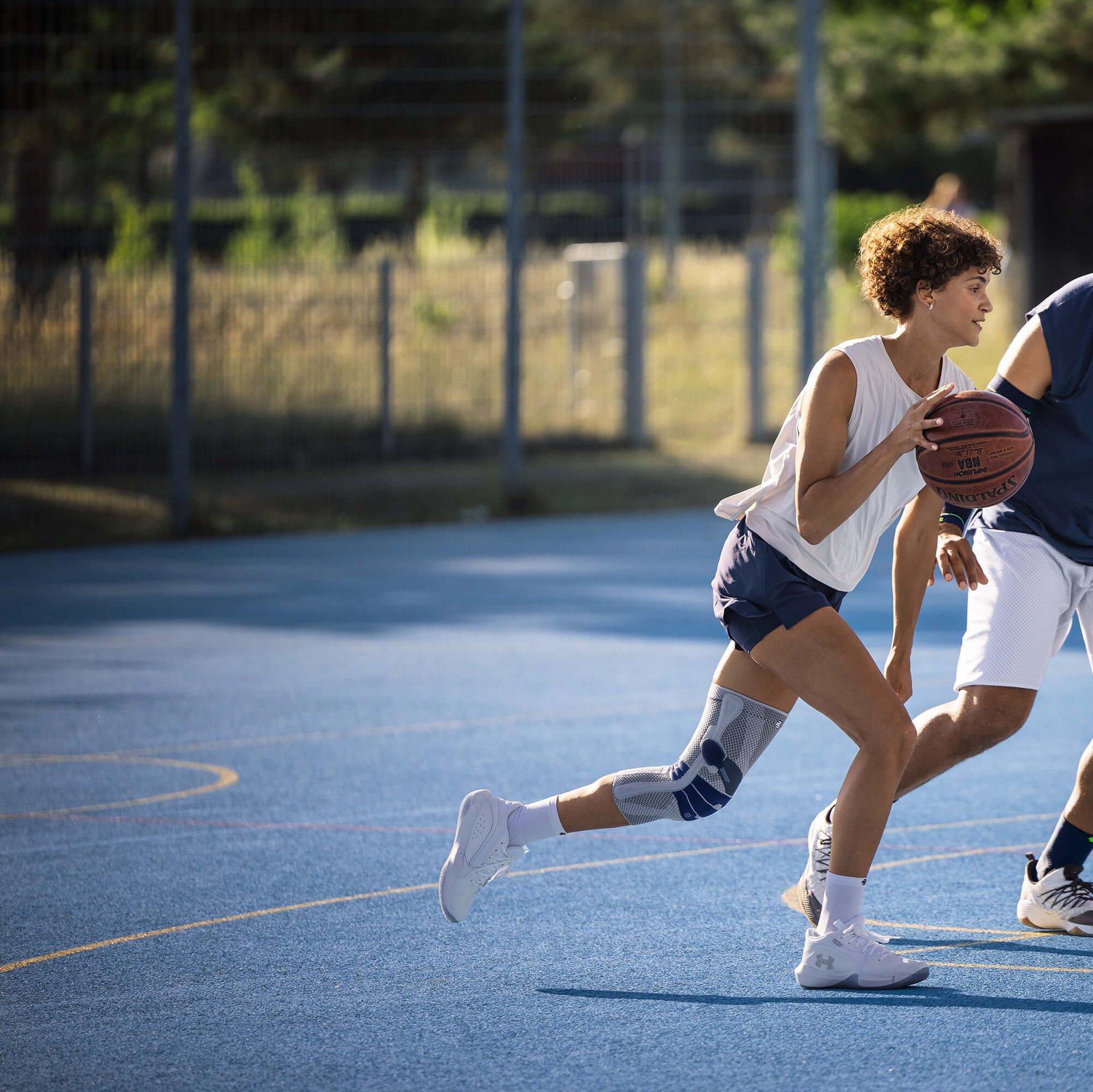Zwei Personen spielen Basketball auf einem Aussenplatz mit blauem Bodenbelag. Die spielende Person im Vordergrund trägt eine Kniebandage am linken Bein und dribbelt den Ball, während die andere Person in Verteidigungsposition vor ihr steht. Beide sind sportlich gekleidet und konzentriert im Spielgeschehen.
