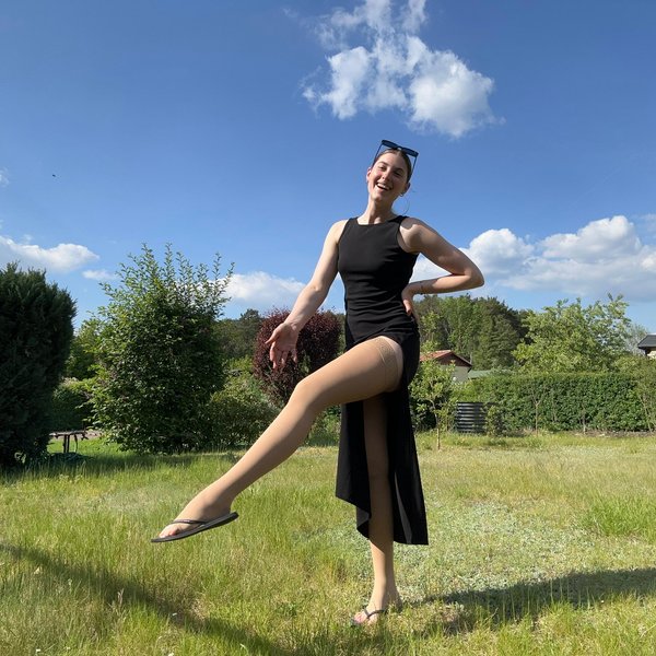 Eva Seemann steht fröhlich in einem Garten und hebt lächelnd ein Bein. Sie trägt ein schwarzes Kleid kombiniert mit Bauerfeind Kompressionsstrümpfen.