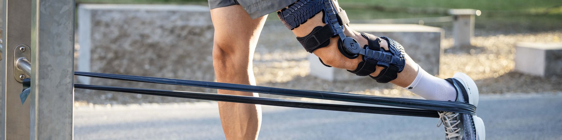 Person trägt graue Sportschuhe, weisse Socken, kurze graue Hose und eine schwarze Knieorthese am rechten Bein; sie führt im Freien eine Übung mit einem Widerstandsband durch, das am Fuss befestigt ist und an einer Metallstange fixiert wurde.