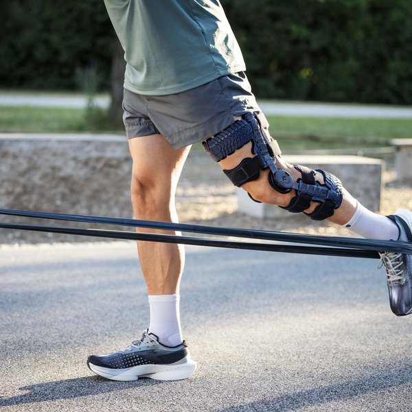 Person trägt graue Sportschuhe, weisse Socken, kurze graue Hose und eine schwarze Knieorthese am rechten Bein; sie führt im Freien eine Übung mit einem Widerstandsband durch, das am Fuss befestigt ist und an einer Metallstange fixiert wurde.