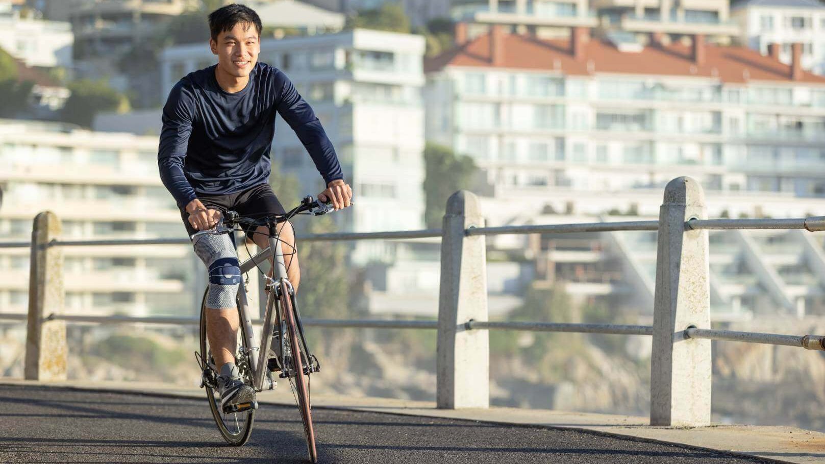 Das Bild zeigt einen Mann, der mit einem Fahrrad an einer Strandpromenade entlangfährt und dabei eine GenuTrain Bandage trägt, um seine Kniebeschwerden zu lindern. Radfahren erweist sich als gelenkschonende Sportart.