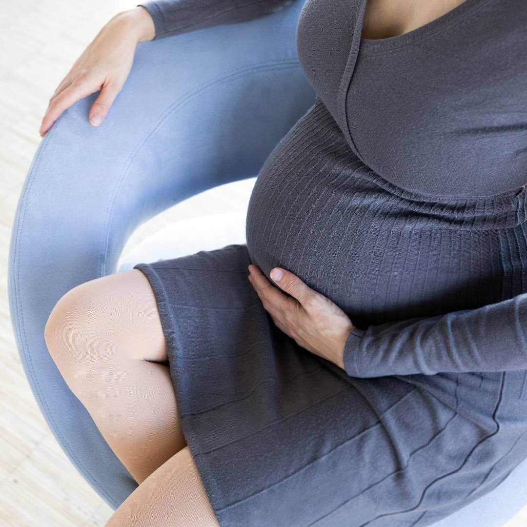Eine schwangere Frau im weißen Kleid steht in einem Hof und lächelt freundlich in die Kamera. Sie trägt einen Cardigan, welcher im Winde weht. Außerdem hat sie eine VenoTrain® micro Schwangerschaftsstrumpfhose von Bauerfeind an, welche die werdende Mutter optimal versorgt.