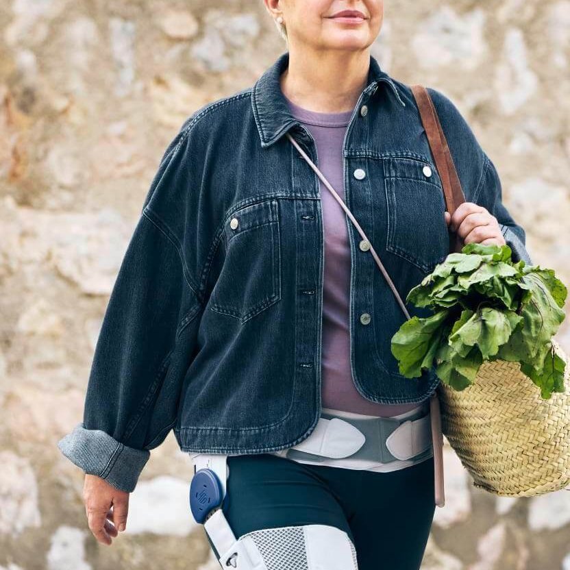 Eine ältere Frau mit kurzen weissen Haaren und Brille überquert einen Zebrastreifen. Sie trägt eine Jeansjacke, ein rosafarbenes Oberteil und dunkle Leggings, an denen medizinische Geräte befestigt sind. In ihrer Hand hält sie einen Korb mit frischem Grünzeug. Im Hintergrund ist eine Steinmauer zu sehen.