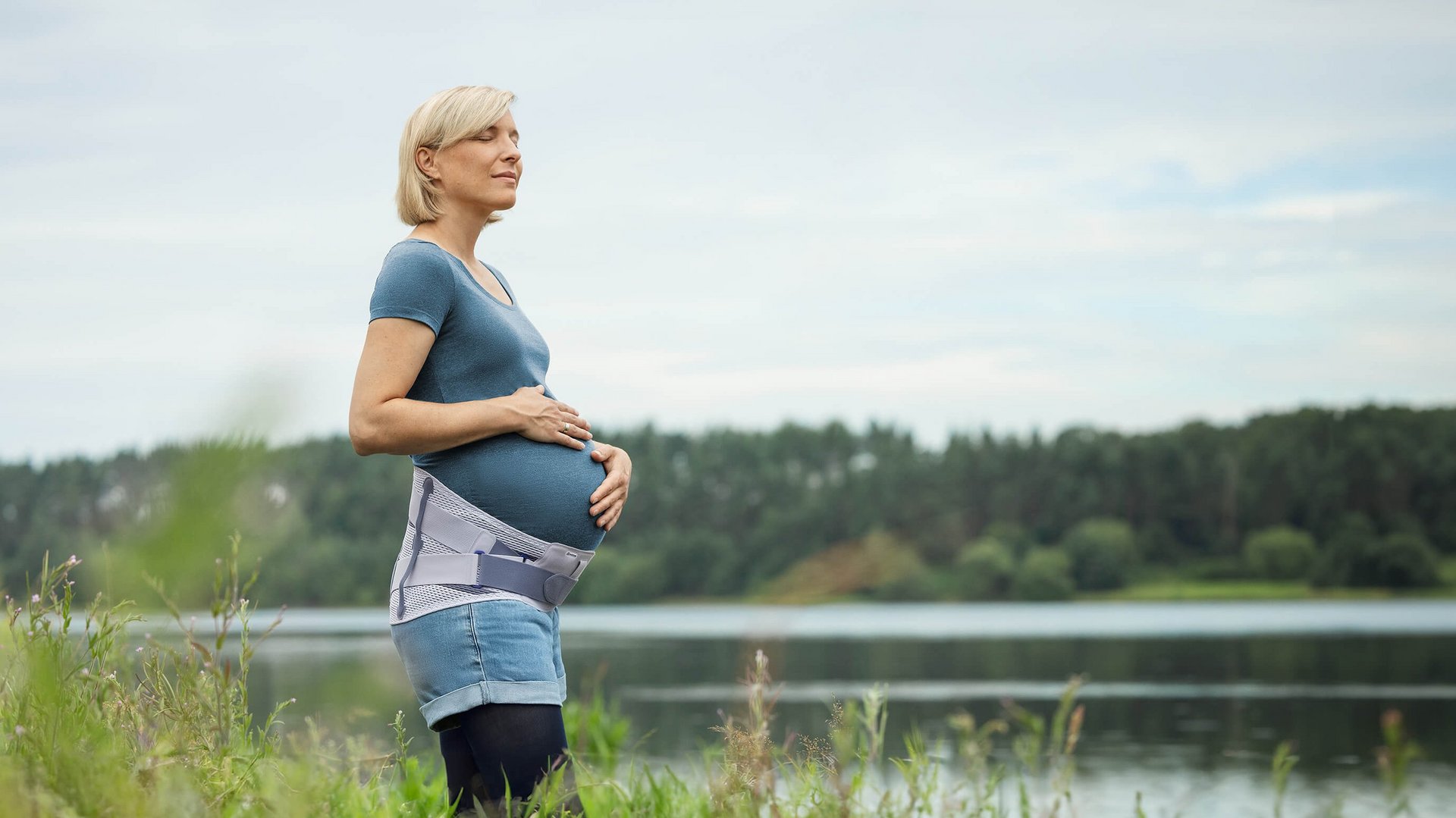 Schwangere Frau steht am Ufer eines Sees und hält ihren Babybauch, während sie in die Ferne blickt. Sie trägt die Spinova Mum Schwangerschaftsorthese und genießt die ruhige Naturkulisse.
