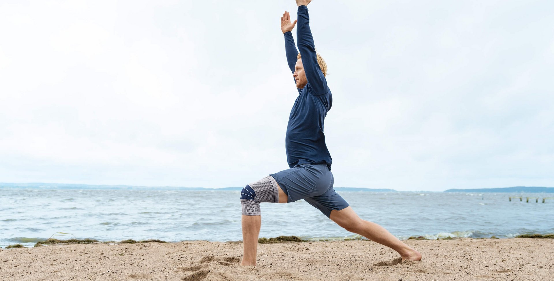 Das Bild zeigt einen Mann in Yoga-Pose am Strand. Am rechten Knie trägt er eine GenuTrain zur Unterstützung. Die Bandage bietet u.a. Unterstützung bei einem Kniegelenkserguss.