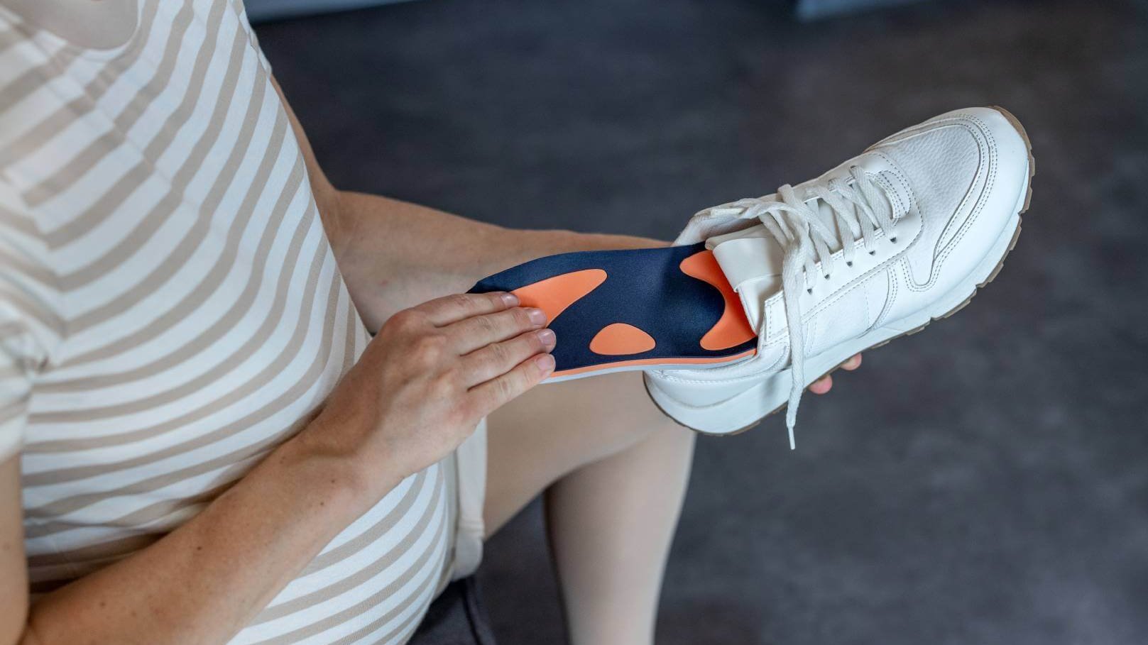 Schwangere Frau legt eine orange-blaue Schuheinlage von Bauerfeind in einen weißen Sneaker ein. Die Einlage ist speziell auf die Bedürfnisse von Schwangeren abgestimmt, um Komfort und Unterstützung beim Gehen zu bieten. 