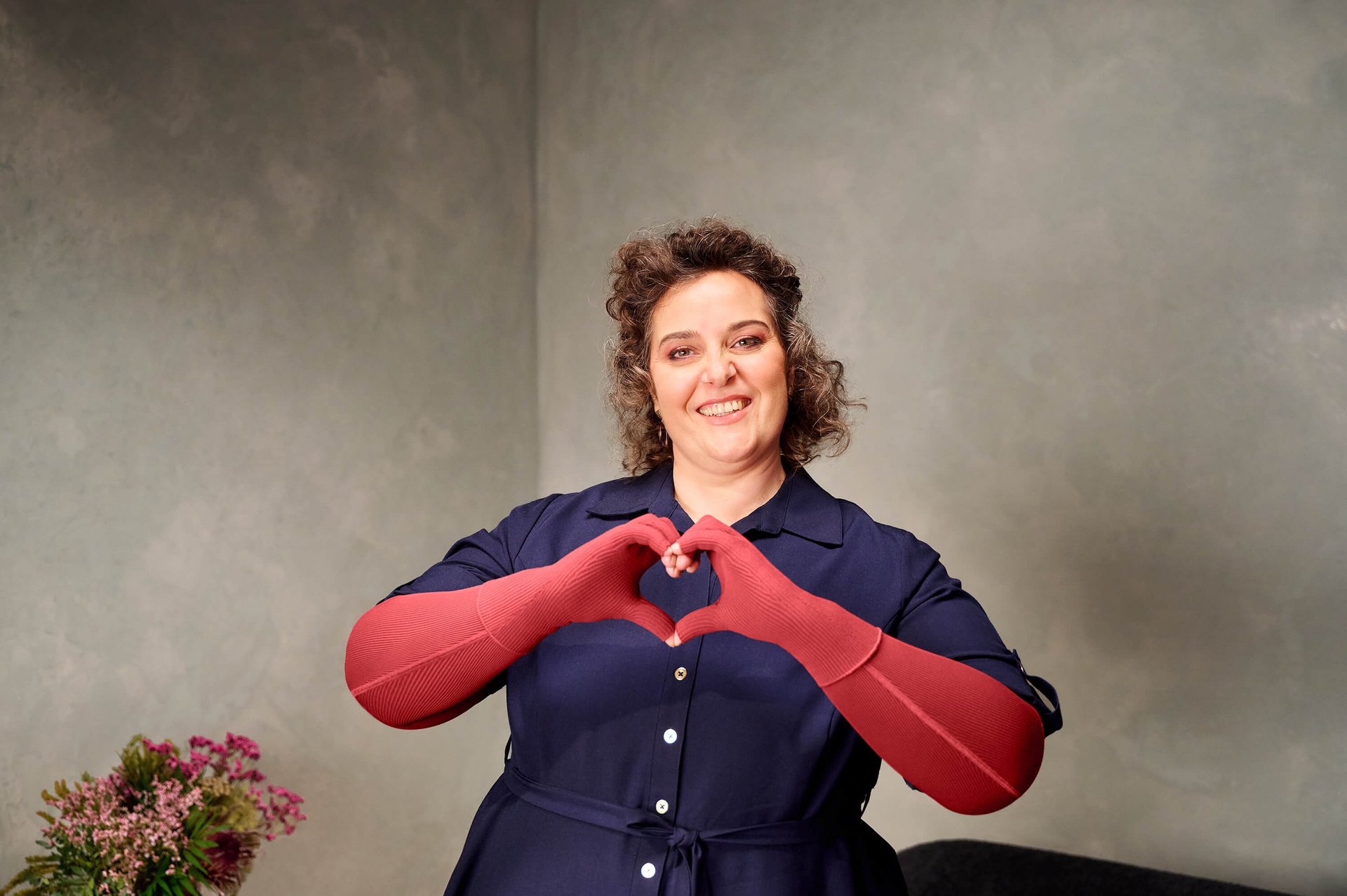 Eine lächelnde Frau steht vor einer hellgrauen Wand und formt mit ihren roten Kompressionshandschuhen ein Herz vor ihrer Brust. Links im Bild ist ein Blumenstrauss zu sehen.