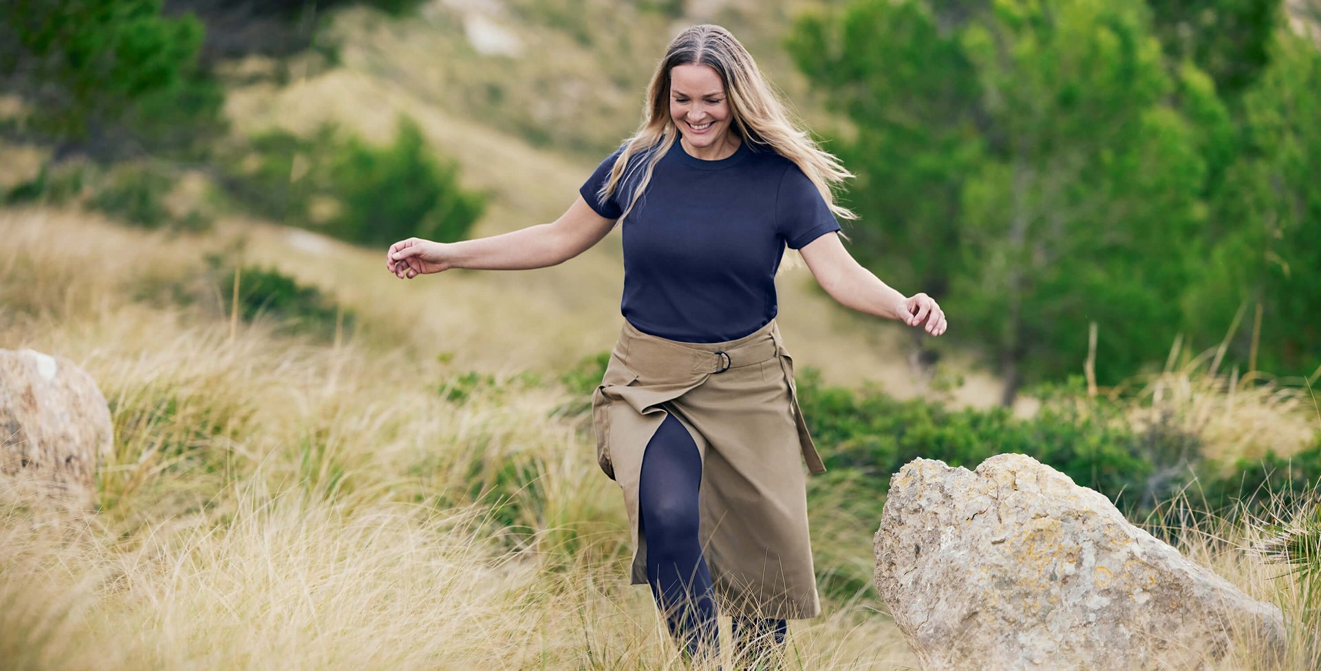Eine lächelnde Frau mit langen, offenen Haaren geht leichtfüßig durch eine hügelige Naturlandschaft mit hohem Gras und Felsen. Sie trägt ein lässiges dunkelblaues T-Shirt, einen khakifarbenen Wickelrock und VenoTrain Kompressionsstrümpfe in Dunkelblau.