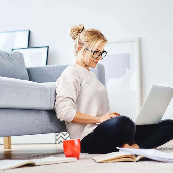 Homeoffice Die Abbildung zeigt eine junge Frau, die mit ihrem Laptop auf dem Boden ihrer Wohnung sitzt und arbeitet.