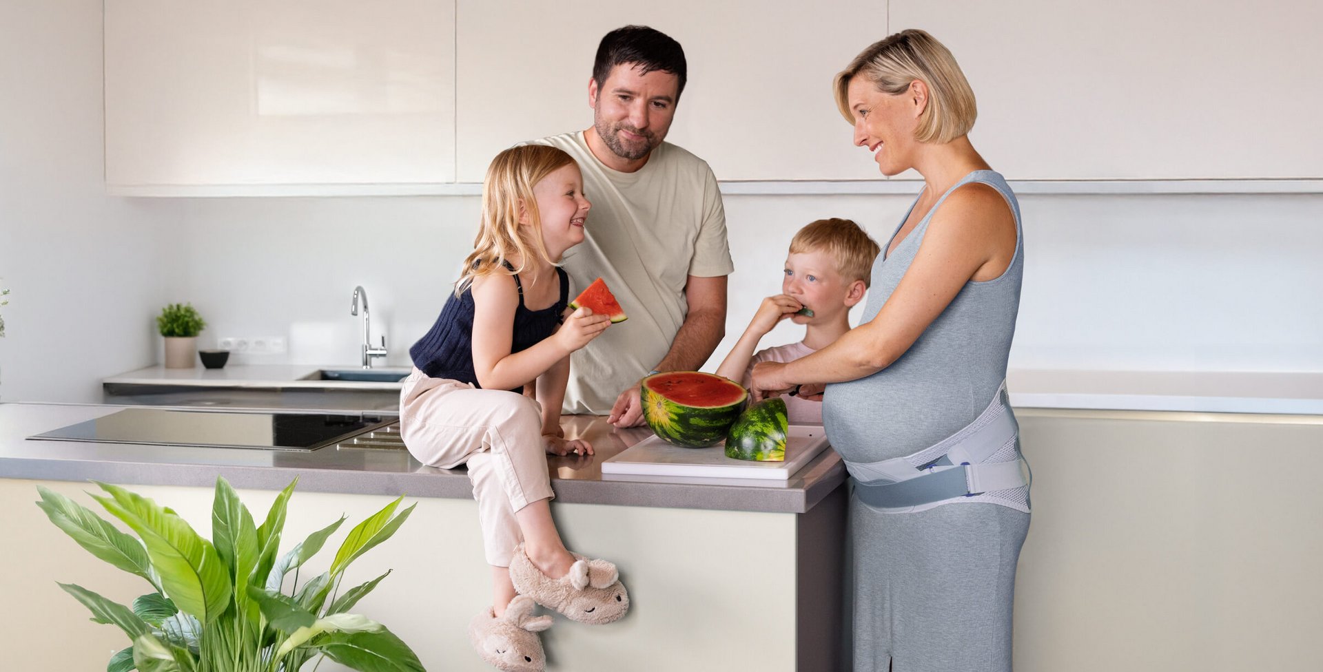 Eine Familie steht in einer modernen Küche um eine Kücheninsel. Ein kleines Mädchen sitzt auf der Arbeitsplatte und hält ein Stück Wassermelone, während ein kleiner Junge ein Stück Melone isst. Ein Mann steht hinter den Kindern, eine schwangere Frau im grauen Kleid mit einer Bauchstütze schneidet die Wassermelone. Alle lächeln sich an.