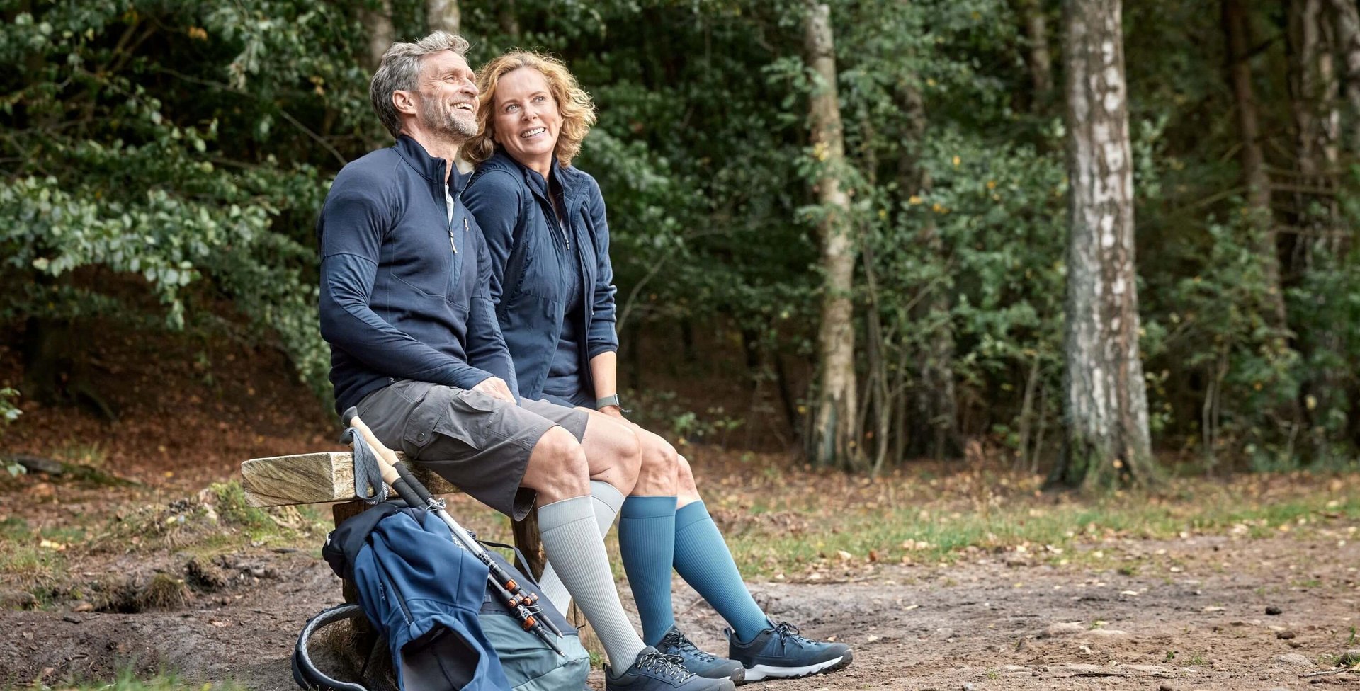 Ein lachendes Paar sitzt auf einer Holzbank im Wald und trägt robuste VenoTrain hero Kompressionsstrümpfe in Grau und Blau. Neben der Bank liegt ein Wanderrucksack mit Wanderstöcken. 
