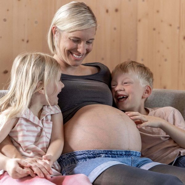  Schwangere Frau sitzt auf einem Sofa und lächelt, während ihre beiden Kinder fröhlich ihren Babybauch berühren.