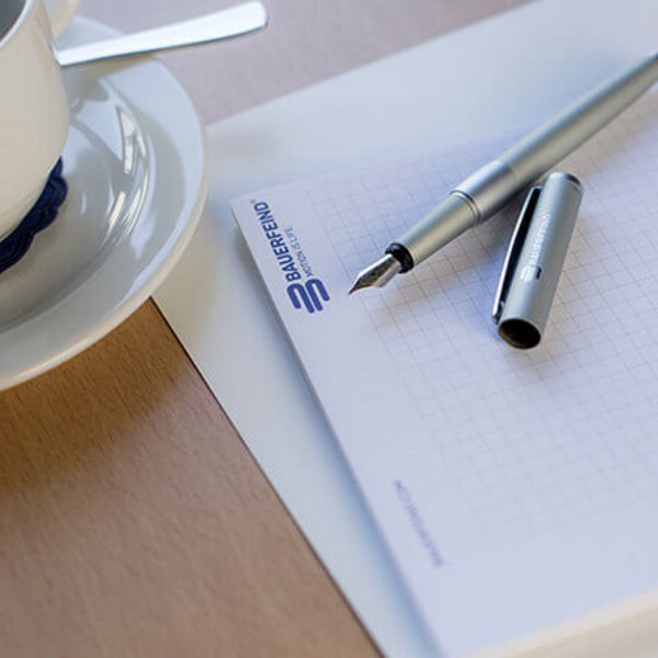 Nahaufnahme eines Notizblocks mit kariertem Papier und dem Logo von Bauerfeind in Blau, daneben ein silberner Füllfederhalter mit abgenommener Kappe. Im Hintergrund eine weisse Tasse mit Untertasse und Löffel auf einem hellen Holztisch.