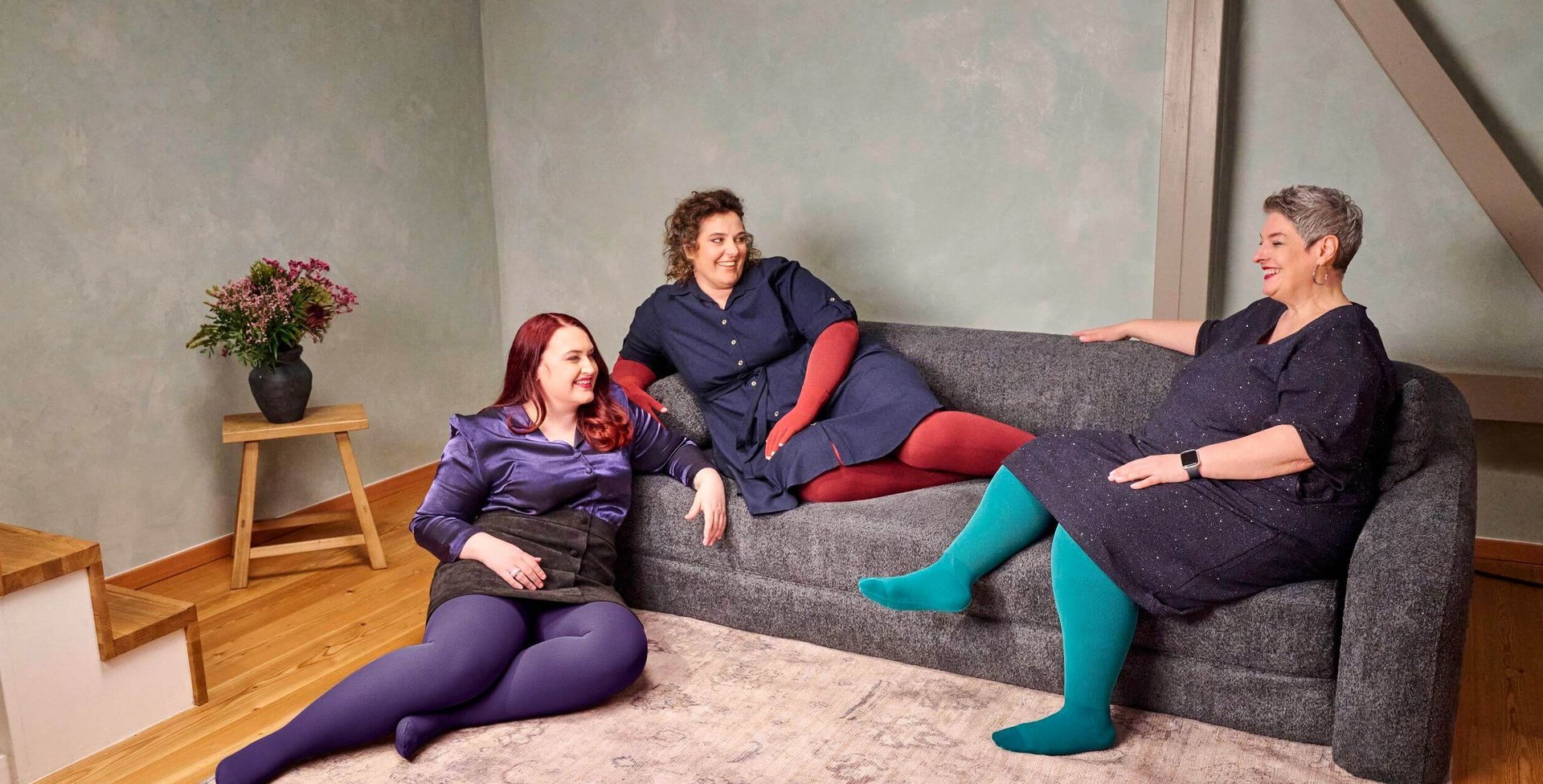 Drei Frauen sitzen und liegen entspannt auf einem dunkelgrauen Sofa in einem modern eingerichteten Wohnzimmer mit Holzfussboden. Sie tragen farbenfrohe Strumpfhosen – in Violett, Rot und Türkis – kombiniert mit dunklen Kleidern oder Röcken. Alle wirken fröhlich und unterhalten sich miteinander. Links steht ein kleiner Holztisch mit einer schwarzen Vase und einem Blumenstrauss.