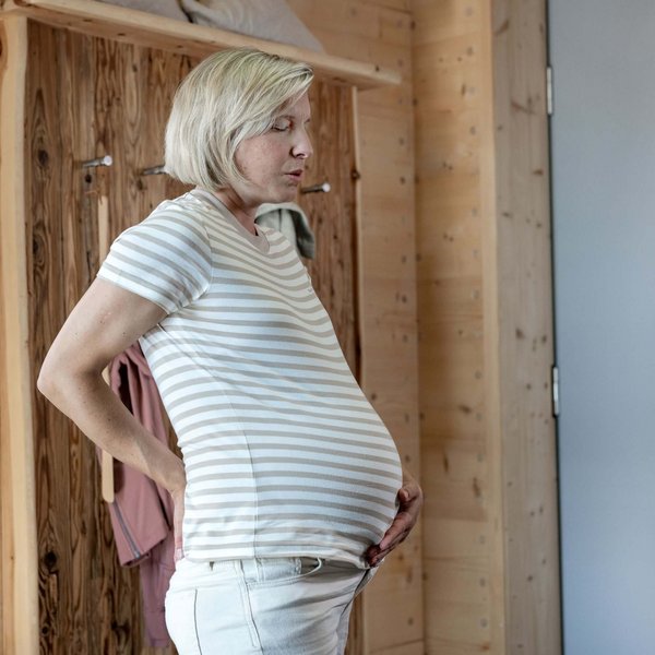 Das Bild zeige eine schwangere Frau, die ihre Hände auf ihren unteren Rücken stützt und tief durchatmet. Die Szene zeigt typische Rückenschmerzen in der Schwangerschaft.