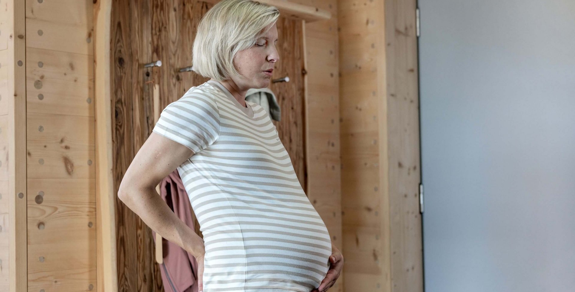 Das Bild zeige eine schwangere Frau, die ihre Hände auf ihren unteren Rücken stützt und tief durchatmet. Die Szene zeigt typische Rückenschmerzen in der Schwangerschaft.