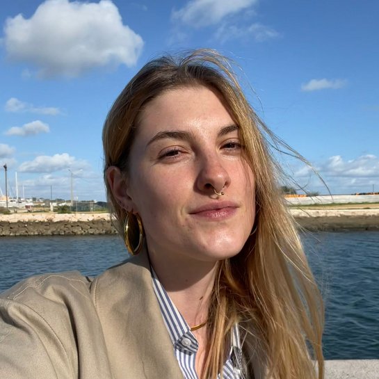 Portrait von Eva Seemann an einem sonnigen Tag am Wasser. Im Hintergrund sind der blaue Himmel, weiße Wolken und ein Hafen mit Gebäuden und Booten zu sehen. 