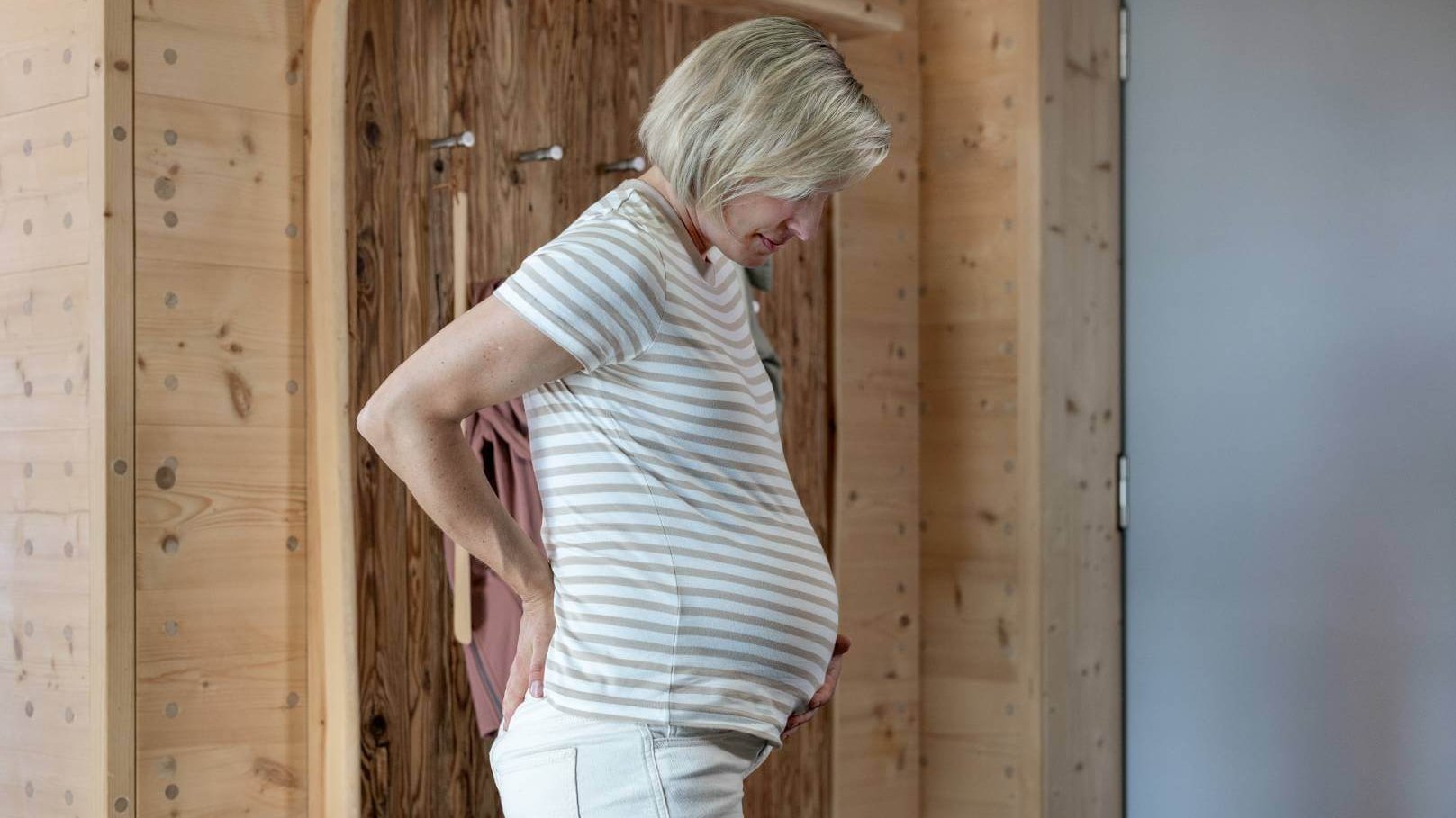 Eine schwangere Frau steht in einem Raum mit Holzwänden und stützt ihre Hände auf ihren schmerzenden unteren Rücken, während sie auf ihren Babybauch schaut. 