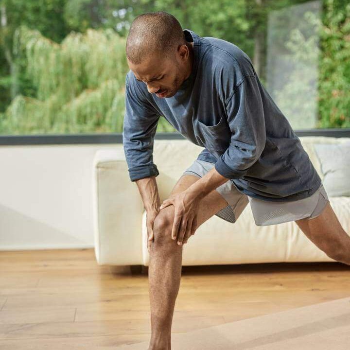 Ein Mann greift sich während des Trainings an das schmerzende Knie. Er trägt sportliche Kleidung und befindet sich in einem modernen Raum mit großen Fenstern. Im Vordergrund sind eine Wasserflasche, ein Springseil und eine Yoga-Matte zu sehen.