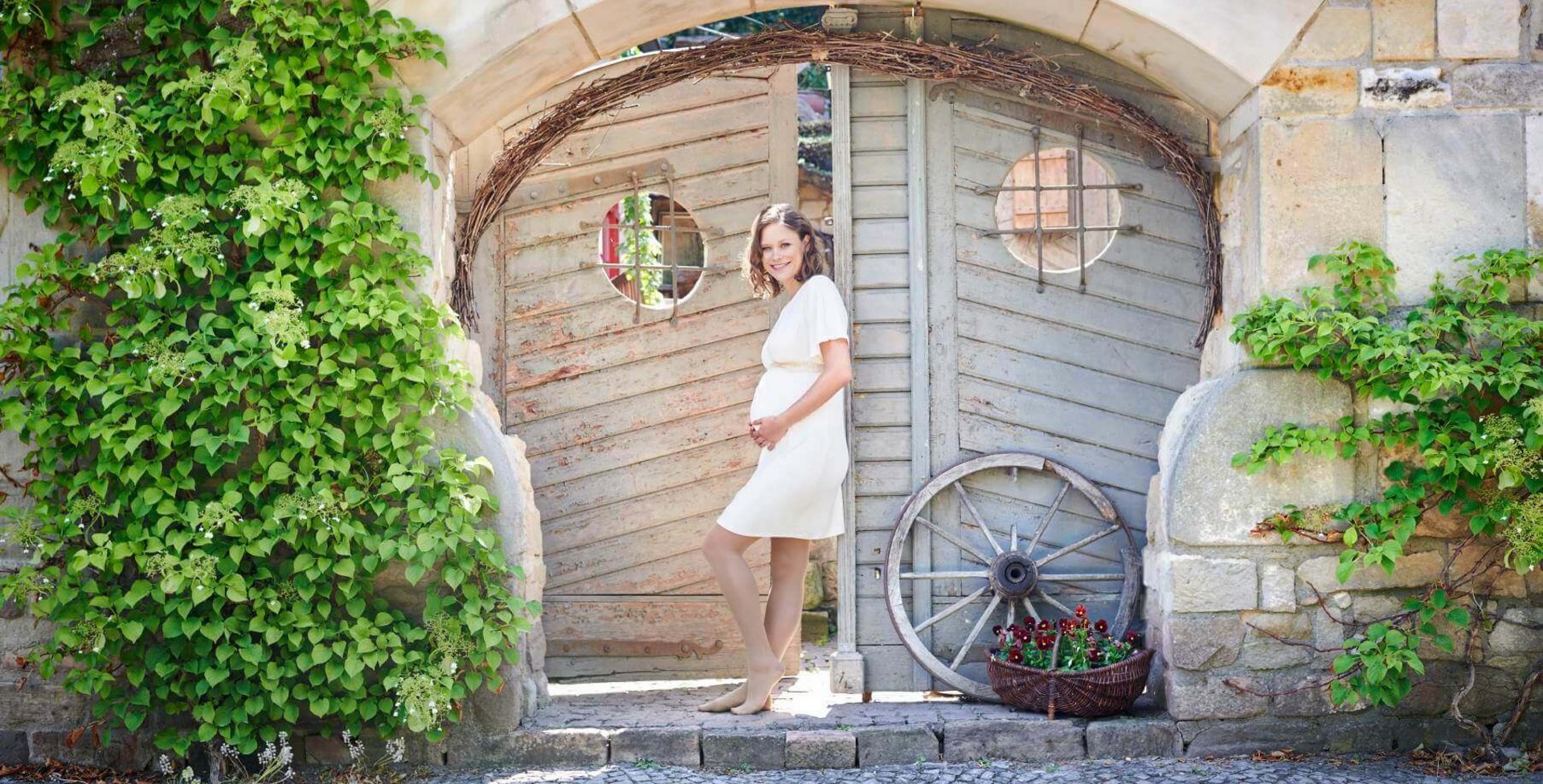 Eine schwangere Frau in einem weißem Kleid steht vor einem Gartentor. Sie hat die Hände auf ihren Babybauch abgelegt und lächelt freundlich in die Kamera. Sie trägt VenoTrain micro Kompressionsstrümpfe von Bauerfeind, welche die werdende Mutter optimal versorgt.