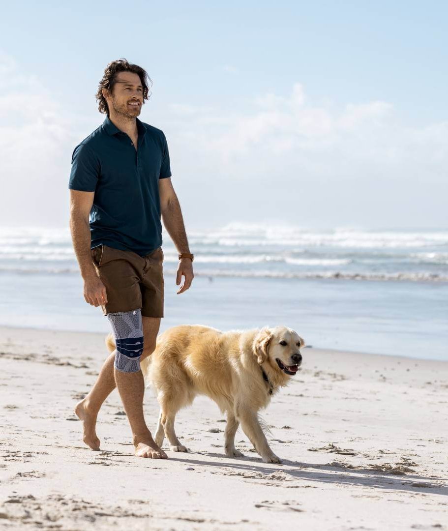 banner-knieschmerzen Ein Mann spaziert barfuß mit einem Golden Retriever an einem sonnigen Sandstrand. Er trägt eine GenuTrain Kniebandage von Bauerfeind, während im Hintergrund Wellen das Ufer erreichen.
