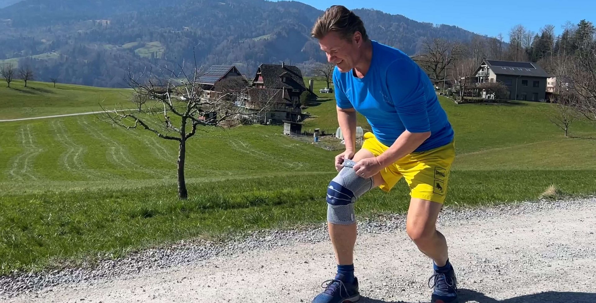 Ein Mann steht auf einem Kiesweg in einer ländlichen Landschaft mit Alpenpanorama im Hintergrund und hält sich das rechte Knie, auf dem eine Kniebandage getragen wird. Er wirkt erschöpft oder hat Schmerzen.  