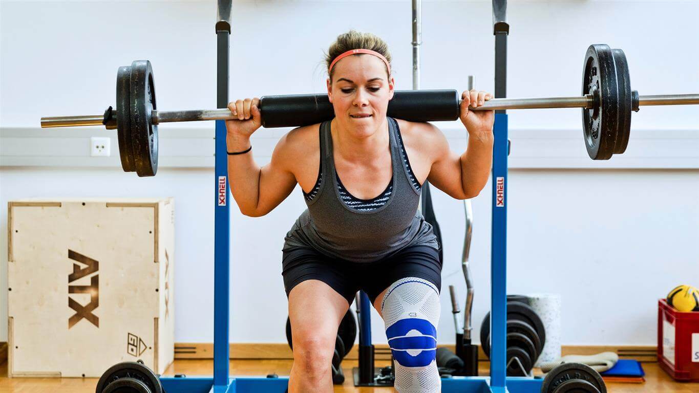 Andrea Limbacher stemmt Gewichte in einem Fitnessstudio. Sie trägt eine Bauerfeind GenuTrain Kniebandage am linken Knie, welche das Knie entlastet, stabilisiert und aktiviert.