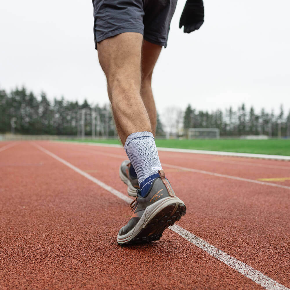 Nahaufnahme der Beine eines Läufers auf einer roten Laufbahn. Der Sportler trägt kurze Hosen, graue Laufschuhe und eine weisse Kompressionssocke mit blauen Details. Im Hintergrund Bäume und ein Sportplatz.