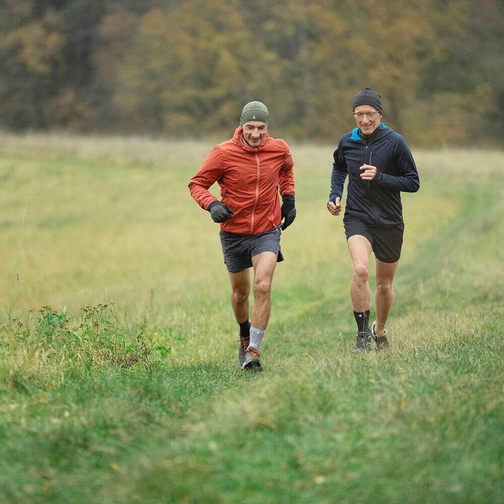Zwei Männer joggen nebeneinander auf einem grasbewachsenen Weg durch eine Wiese. Beide tragen Sportbekleidung mit Mütze und Jacke; einer trägt eine orange Jacke, der andere eine dunkelblaue. Im Hintergrund herbstliche Bäume.