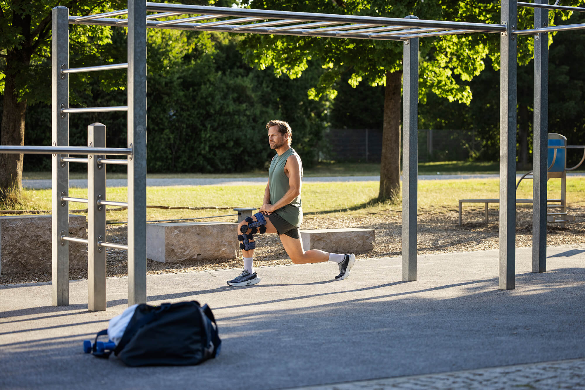 Ein Mann mit Knieorthese führt draussen einen Ausfallschritt unter einer Kletterstruktur aus. Im Vordergrund liegt eine Sporttasche, im Hintergrund sind Bäume und ein Parcours sichtbar.