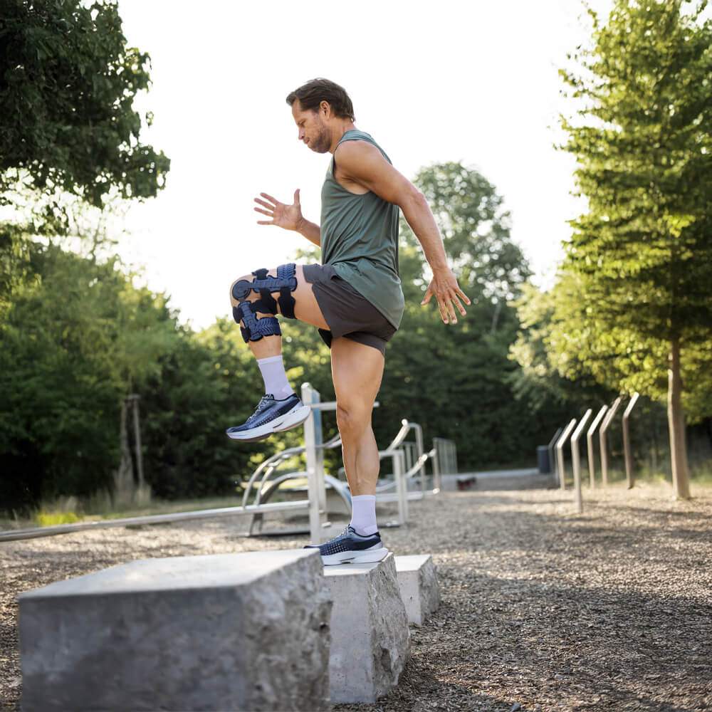 Ein sportlicher Mann mit Knieorthese am rechten Bein macht ein Step-up-Training auf einen grossen Betonblock in einem Outdoor-Fitnesspark. Er trägt ein ärmelloses Shirt, Shorts und Sportschuhe.