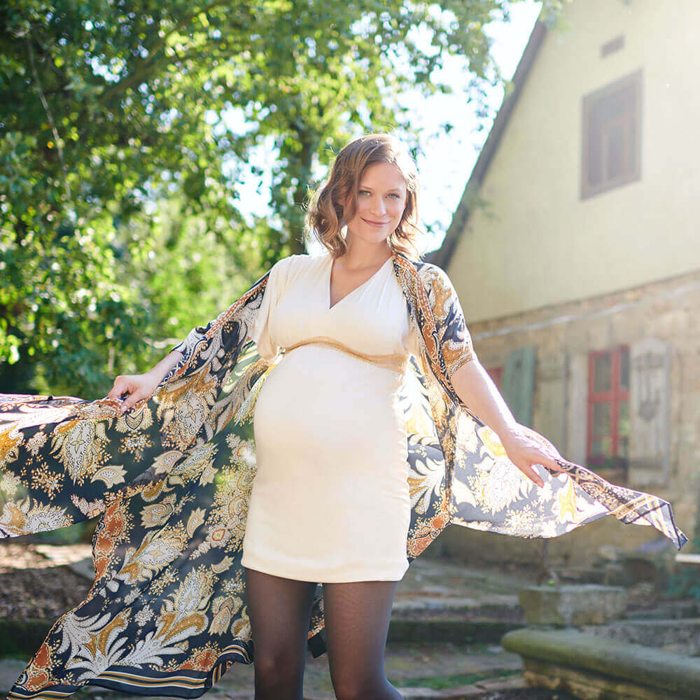 Eine schwangere Frau im weißen Kleid steht in einem Hof und lächelt freundlich in die Kamera. Sie trägt einen Cardigan, welcher im Winde weht. Außerdem hat sie eine VenoTrain® micro Schwangerschaftsstrumpfhose von Bauerfeind an, welche die werdende Mutter optimal versorgt.