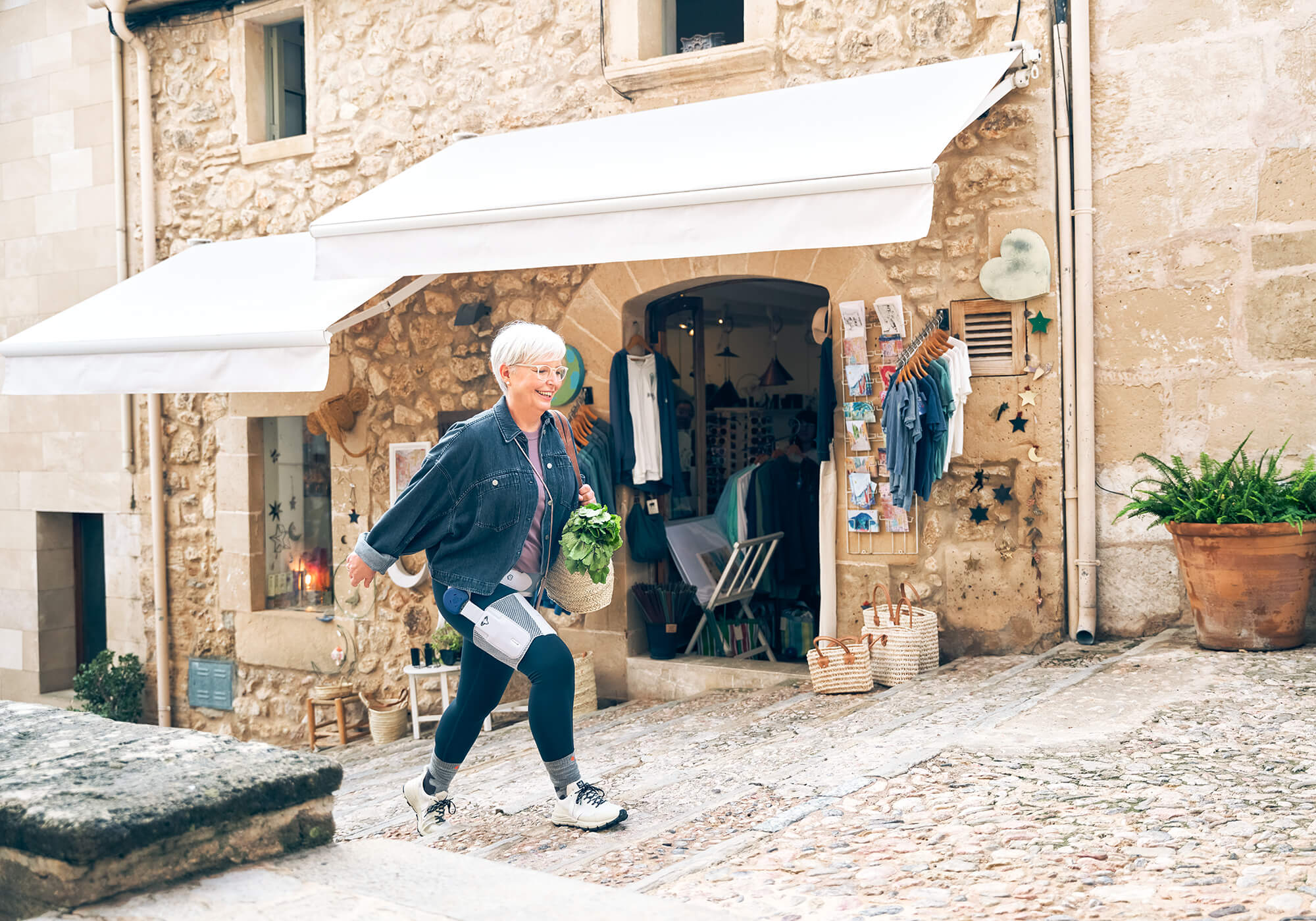 Eine ältere Frau mit kurzen weissen Haaren und Brille läuft eine gepflasterte Gasse hinauf. Sie trägt eine Jeansjacke, dunkle Leggings mit medizinischen Geräten daran und hält einen Korb mit frischem Grünzeug. Im Hintergrund befindet sich ein Geschäft mit Kleidung und Postkarten, eingerahmt von einer Steinwand mit weissen Markisen. Neben dem Eingang stehen Körbe und ein grosser Blumentopf.