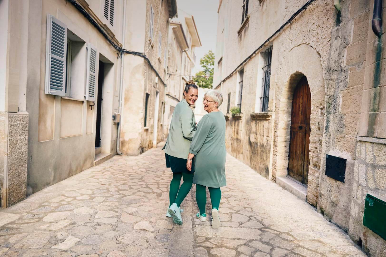 Zwei Frauen, eine jüngere und eine ältere, spazieren lachend Hand in Hand durch eine charmante, enge Altstadtgasse. Beide tragen farblich abgestimmte Outfits in Grün mit passender Kompression und Sneakern.