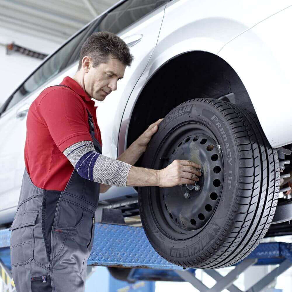 Das Bild zeigt einen Mechaniker in Arbeitskleidung, der einen Autoreifen montiert.Dabei trägt er eine EpiTrain Ellenbogenbandage von Bauerfeind, die bei einem Golferellenbogen Unterstützung und Schmerzlinderung bietet.