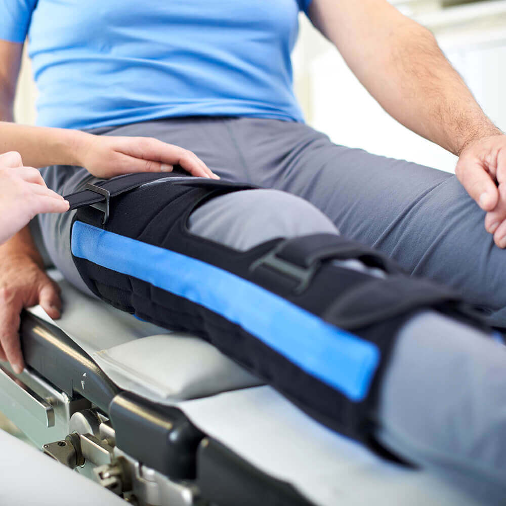 Bildausschnitt eines Mannes auf einer Therapieliege. Zur Ruhigstellung seines verletzten Knies trägt er eine GenuLoc Orthese von Bauerfeind.