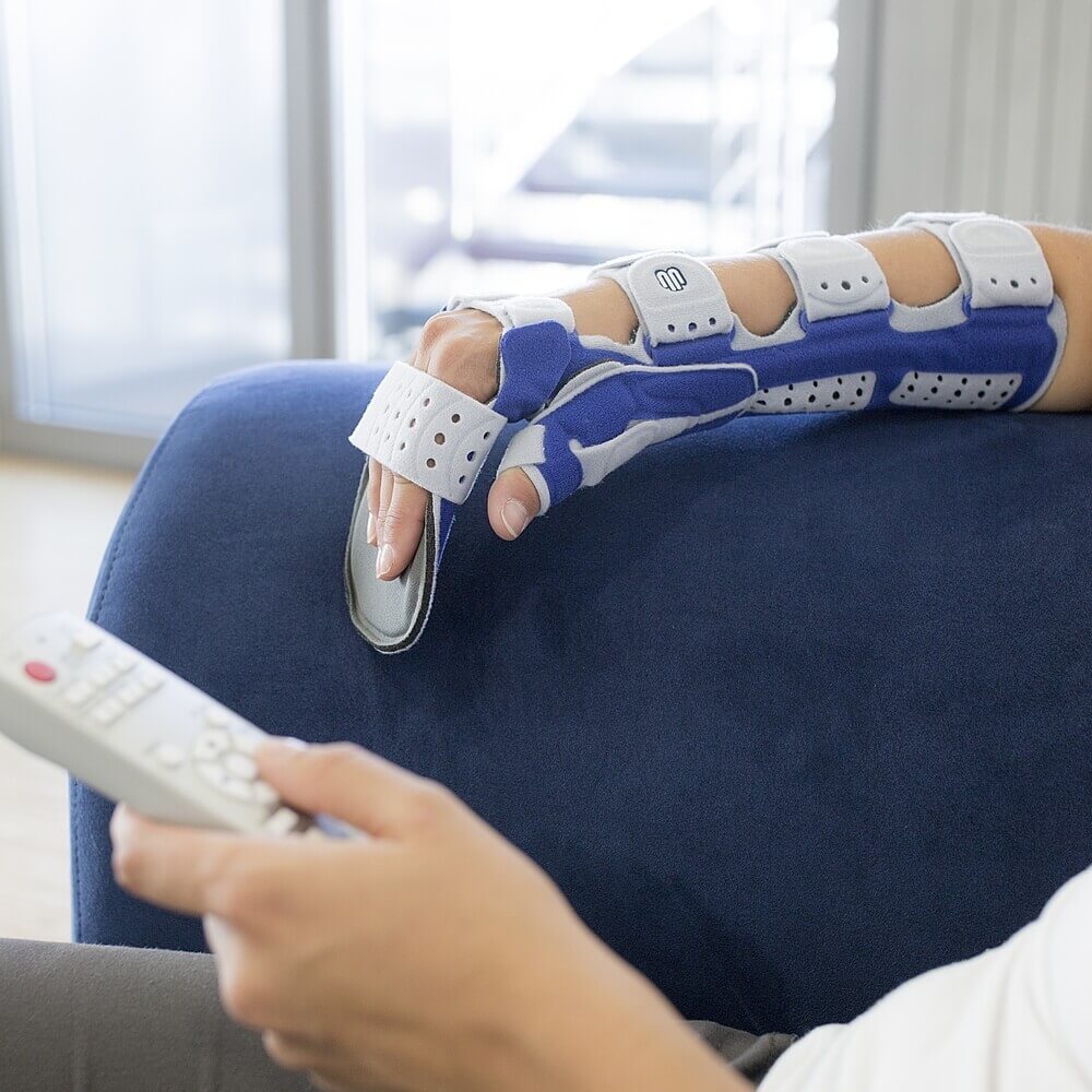Bildausschnitt einer Frau mit Fernbedienung auf einem blauen Sofa. An ihrer rechten Hand trägt sie eine ManuLoc Long Plus Orthese von Bauerfeind.